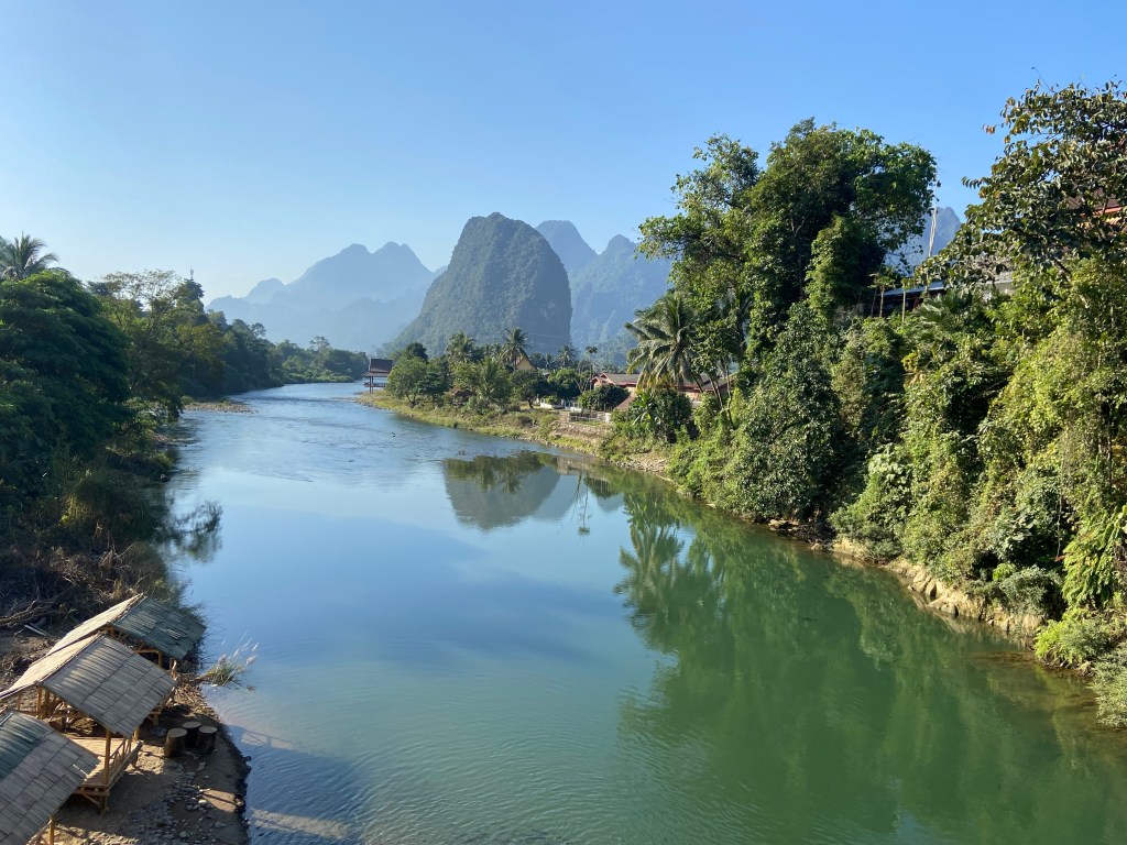 Wide and slow moving river with forested mogotes in the background on the right side of the river and jungle on the left. In the foreground on the river edge there are bamboo huts