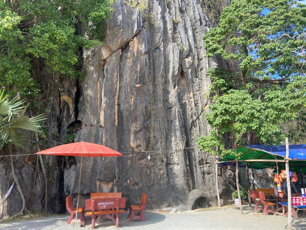 Small grey limestone crag with a food stall, benches and tables set up at the base. They have wires to support the stall and lights hanging from the first bolts of what would be the sport climbs. 