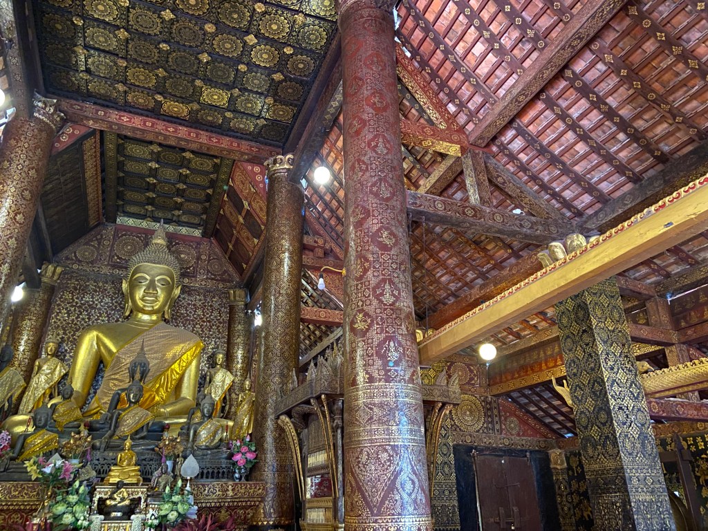 Large golden Buddha statue surrounded with smaller bronze and golden buddhas. They sit within an ornately decorated temple with patterned pillars and roof.