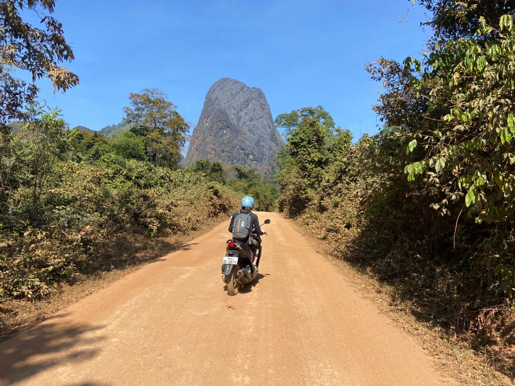 Woman riding motorbike with blue helmet and backpack on dirt road heading towards a tiny but tall limestone peak 