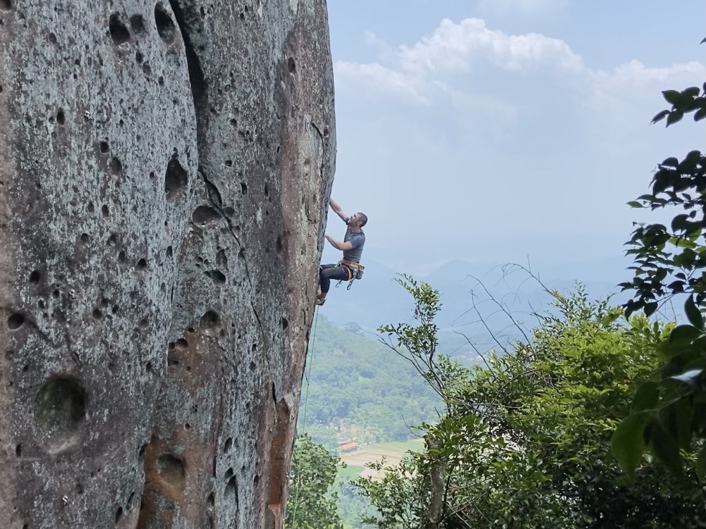 Climber on a slightly overhanging wall looking for the next move. The rock is pocketed pink and grey andesite. Below are the flat green rice fields and small villages. Behind these are more jungle covered mountains in the haze. 