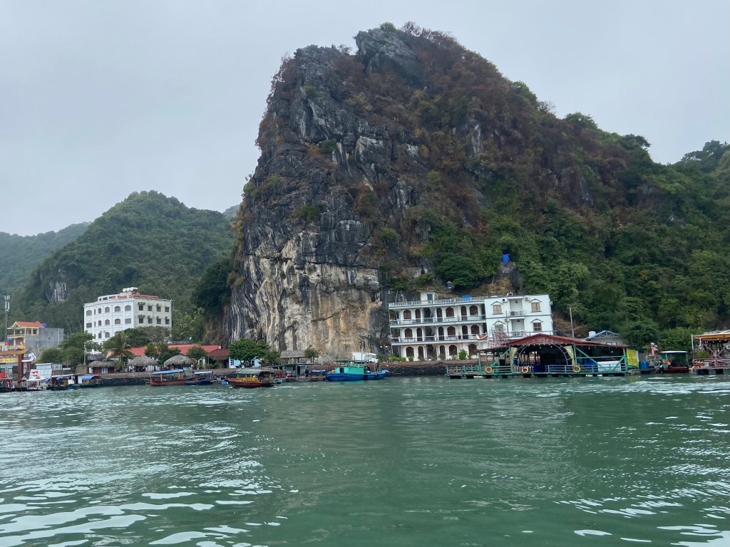 On the water looking towards the harbour which is filled with docked boats. There are some large buildings and a grey black limestone crag which has some climbing routes on. 