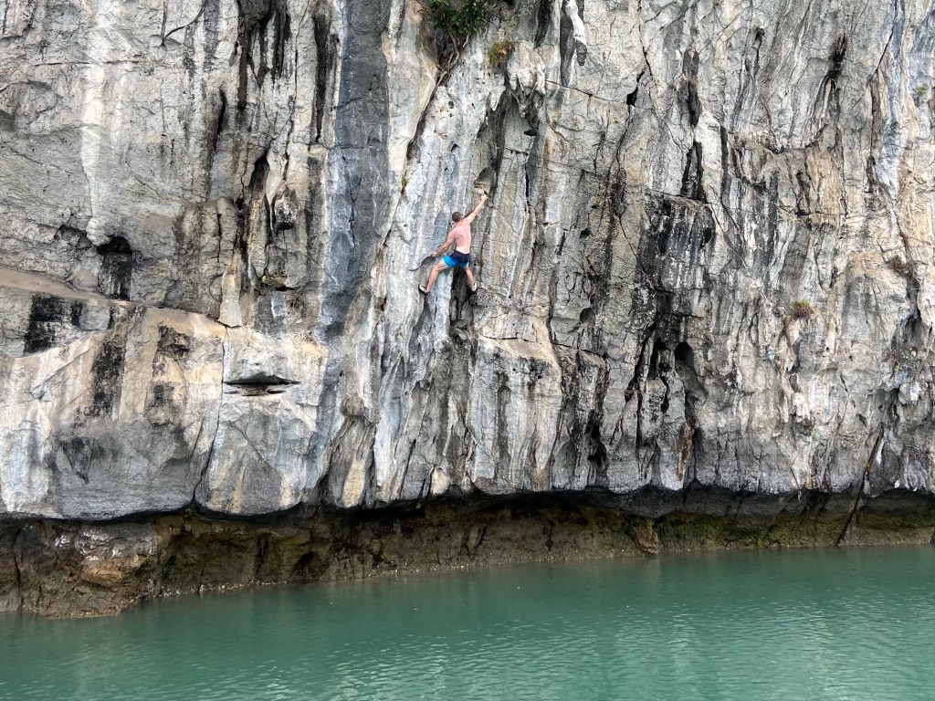 Climber reaching out to a high hand hold high above the sea on a slightly overhanging white limestone cliff with grey and black streaks. 