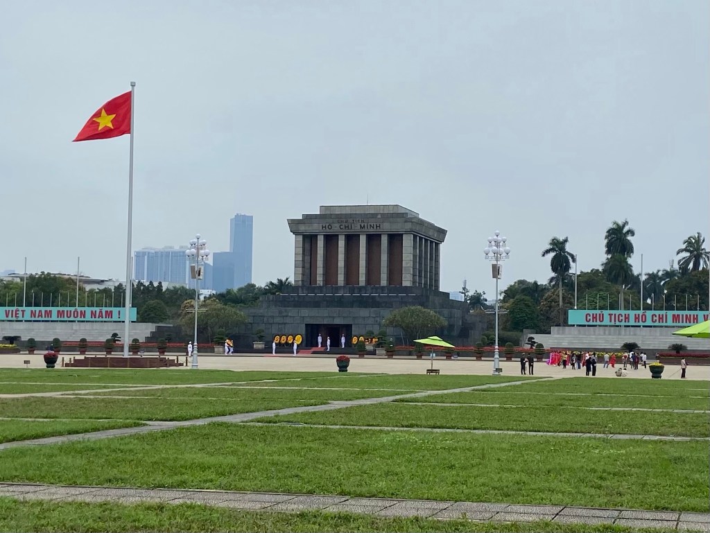 The large brutalist style mausoleum of Ho Chi Minh. In front are guards dressed in a white uniform and a large flag of Vietnam flying. Behind the building are some modern skyscrapers of Hanoi. 