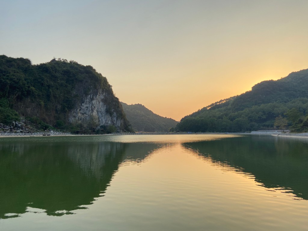 Sunset turning the skying an orange colour at a reservoir at Fisher Valley. Behind the reservoir is the cliff of Fisher Valley and some tree covered hills which have a reflection on the water.