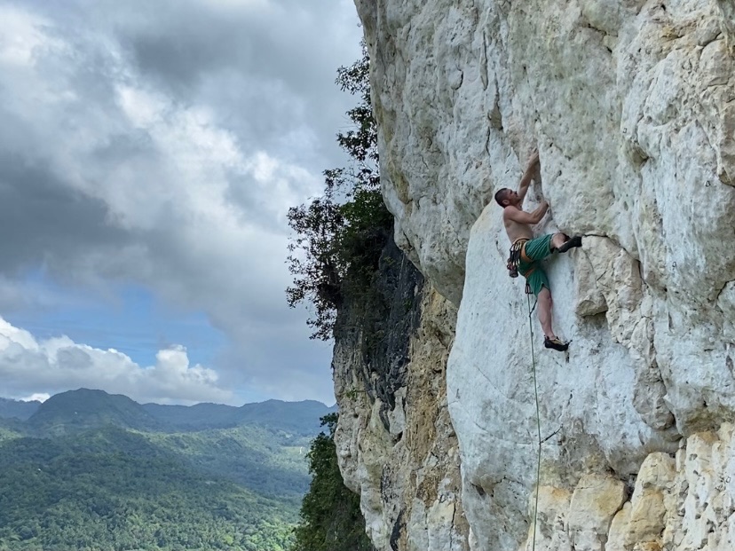 Climber trying hard on a thin section of vertical white limestone. In the background are mountains and jungle. 