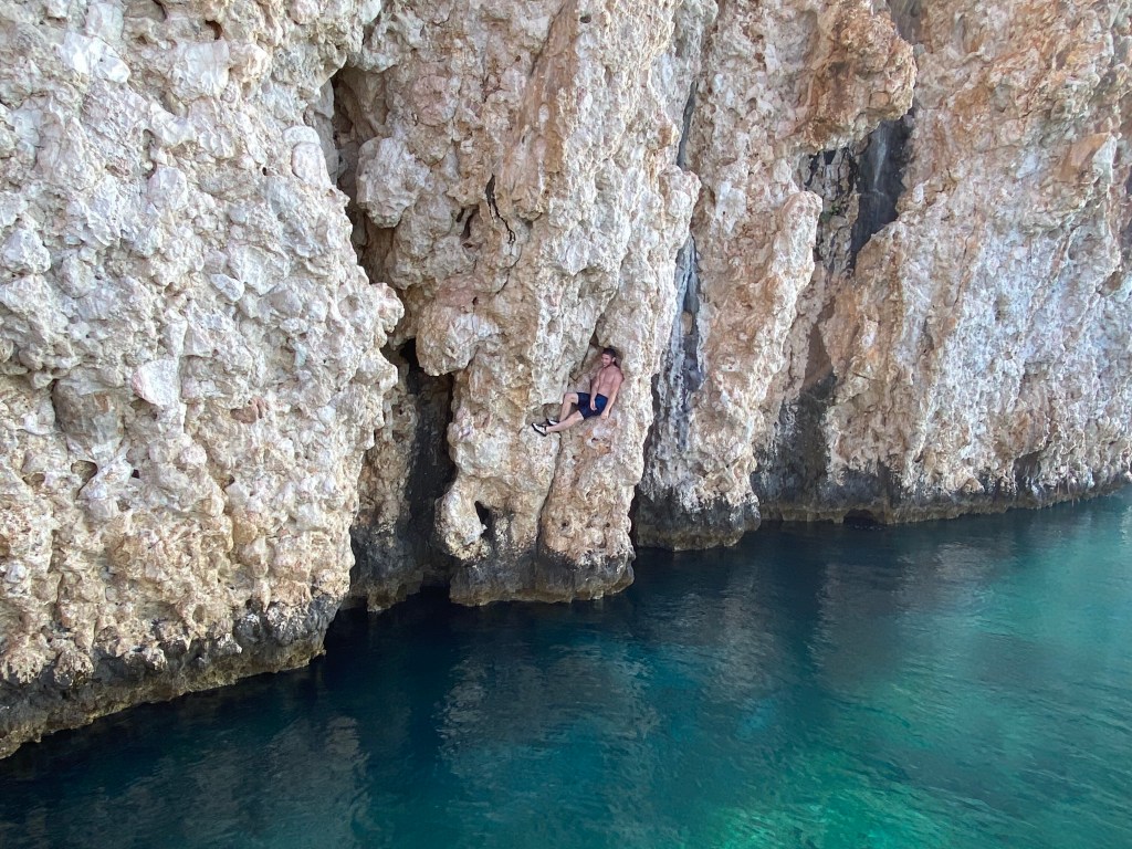 Climber using a depression in the rock to use a no hands rest, sitting down a few meters above the water. 
