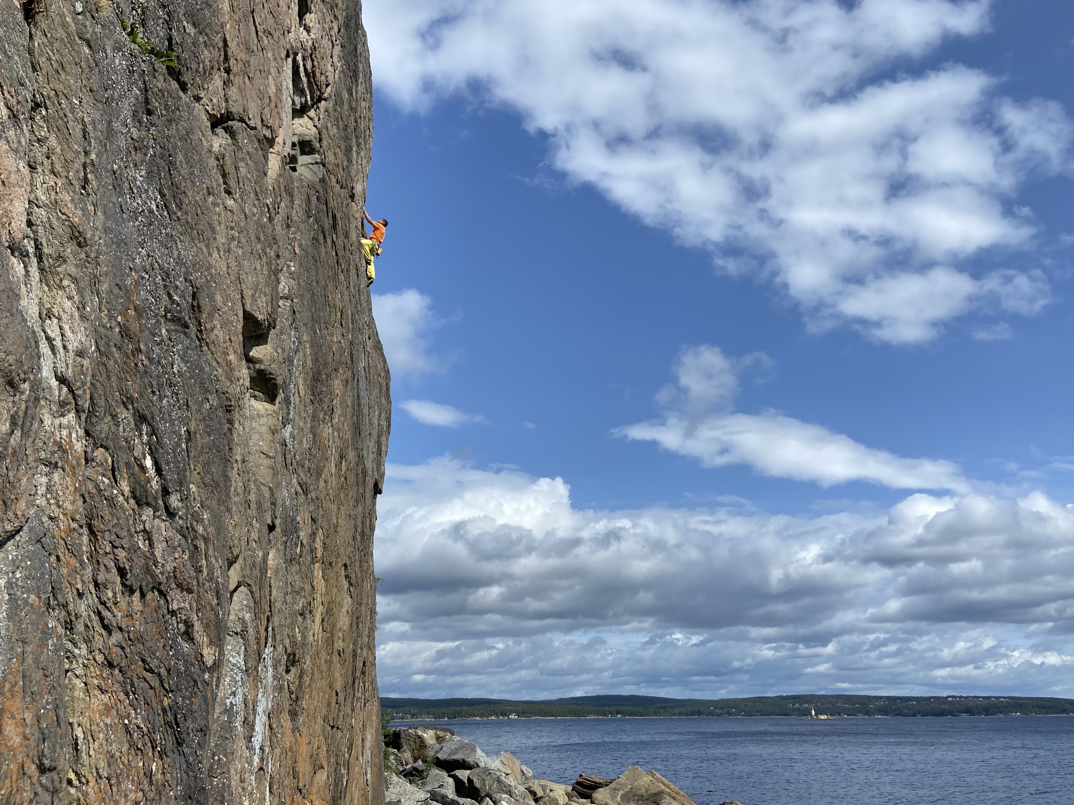 Climber in an orange t-shirt and yellow trousers on an exposed aerate climb above the rocky beach and sea at Brattberget, Sundvall.