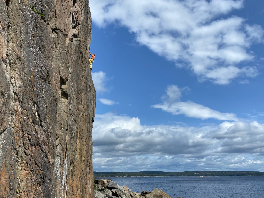 Climber in an orange t-shirt and yellow trousers on an exposed aerate climb above the rocky beach and sea at Brattberget, Sundvall.