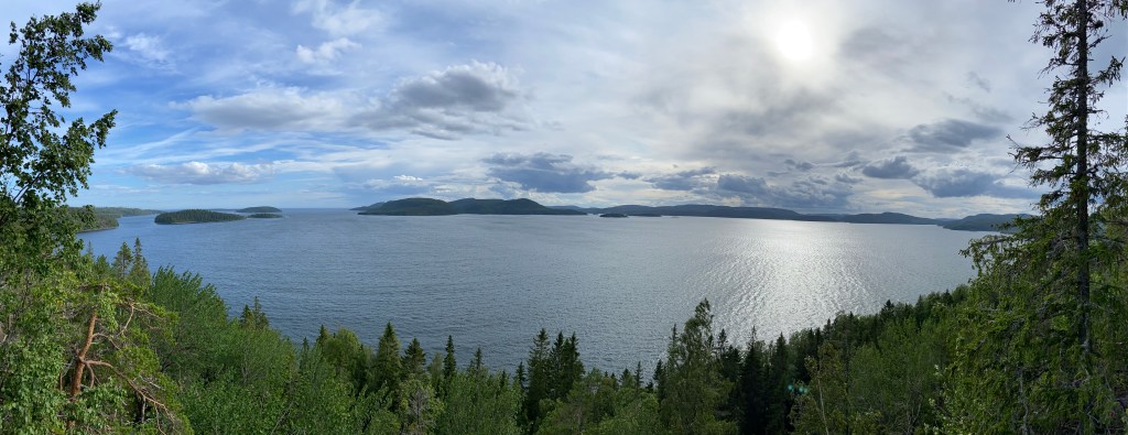 Steep pine forest down to the calm inlet of the sea with small forested islands and peninsulas in the background and wispy clouds in the sky