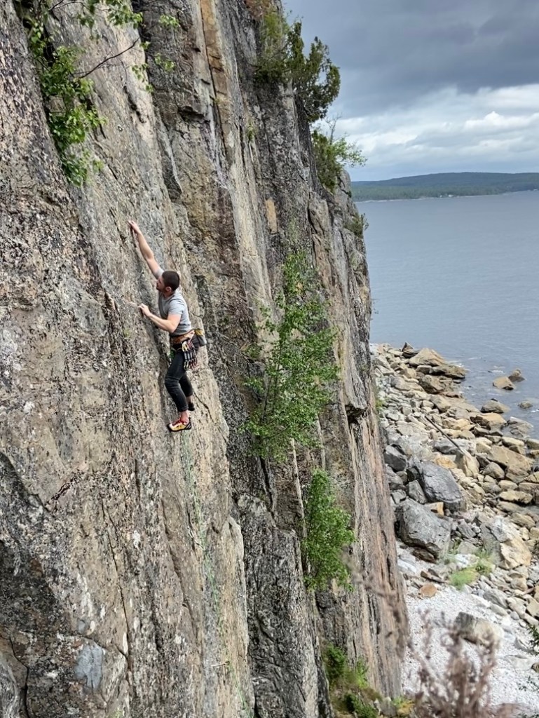 Climber reaching for high crimps on the grey compact slab above the rocky beach 