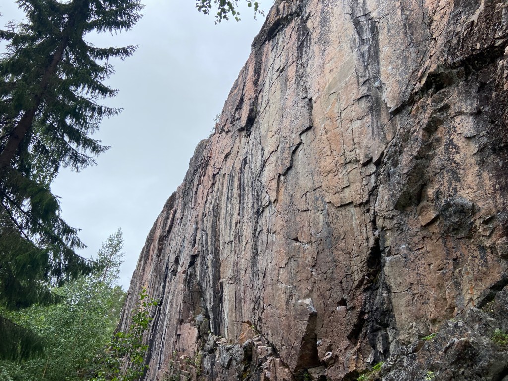 Tall and striking pink granite cliff face with black streaks. There are vertical, and rising cracks as well as sharp undercut features. 