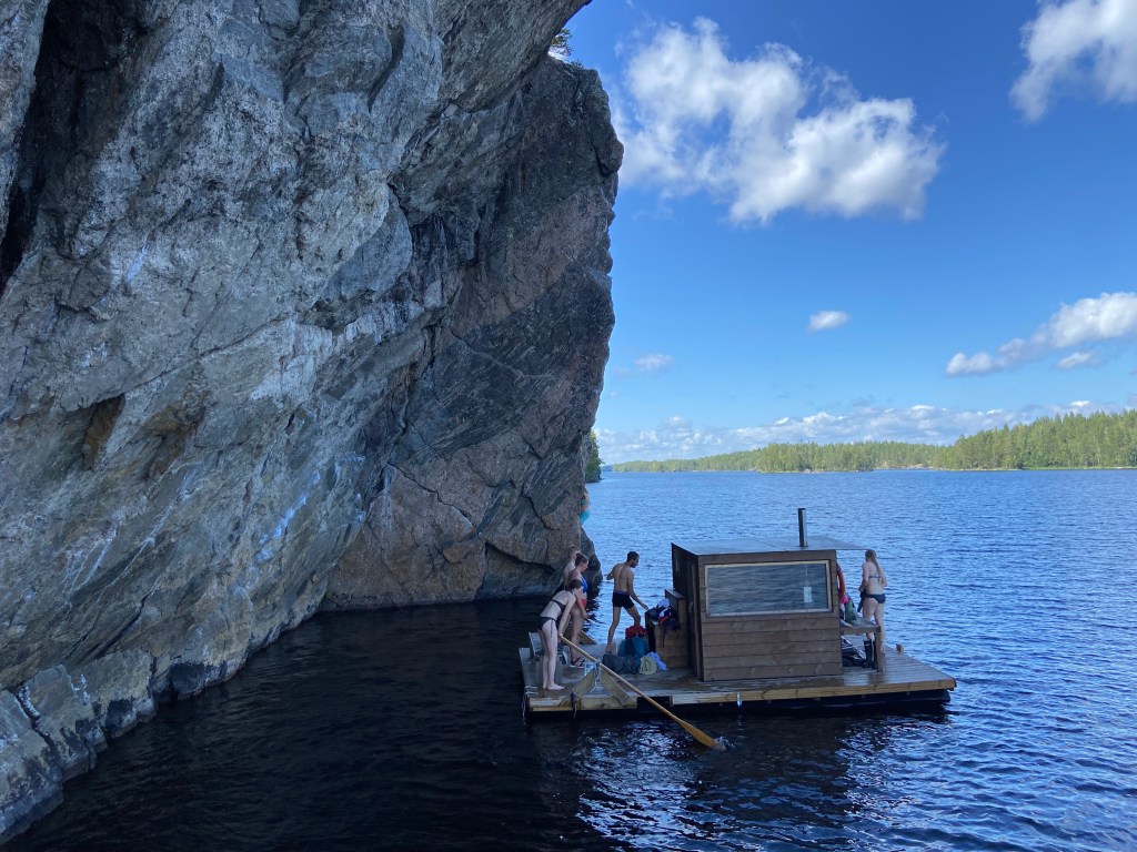 People steering the floating platform with a sauna in the middle below the steep granite cliff on a lake. The far side of the shore is lined with trees  