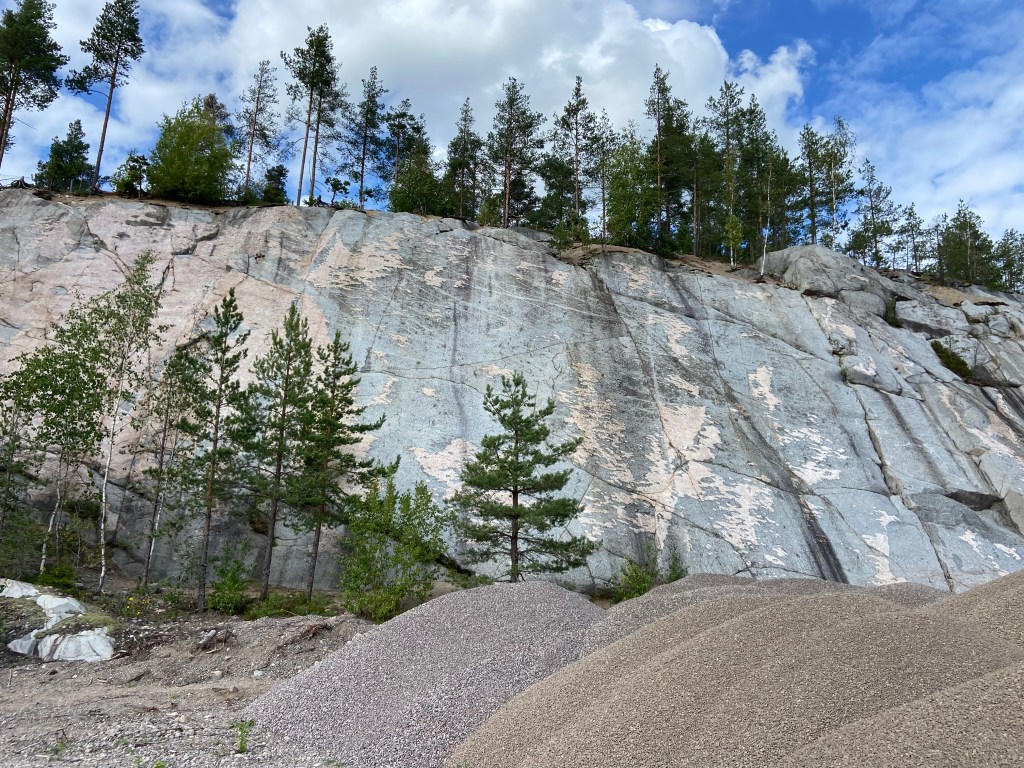 Super slabby piece of granite with grey and pink features. There are trees at the base and at the top. In front of the crag are piles of gravel from the building yard.