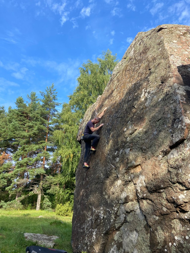 Climber using a high foothold to make their way up the tall boulder with the pine forest in the background