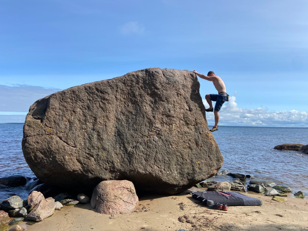 Climber padding their feet up the arete on a smaller boulder on the beach at the edge of the sea