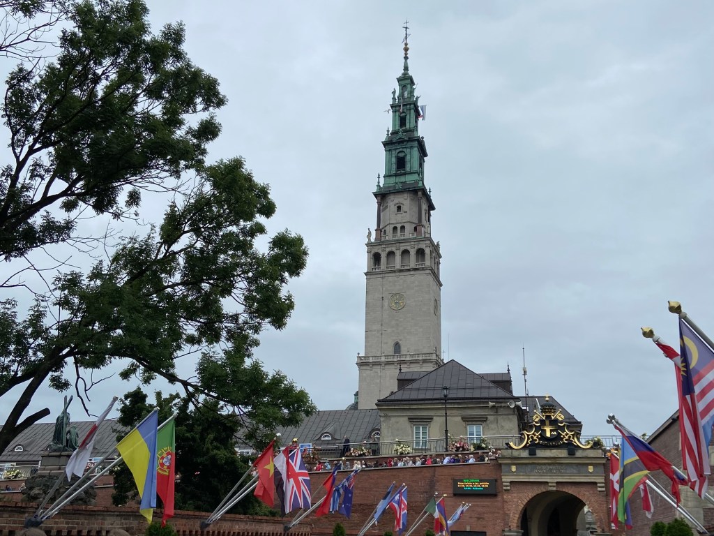 Tall church spiral behind a brick wall and archway protecting the monastery complex. The entry way is lined with many different flags from around the world.