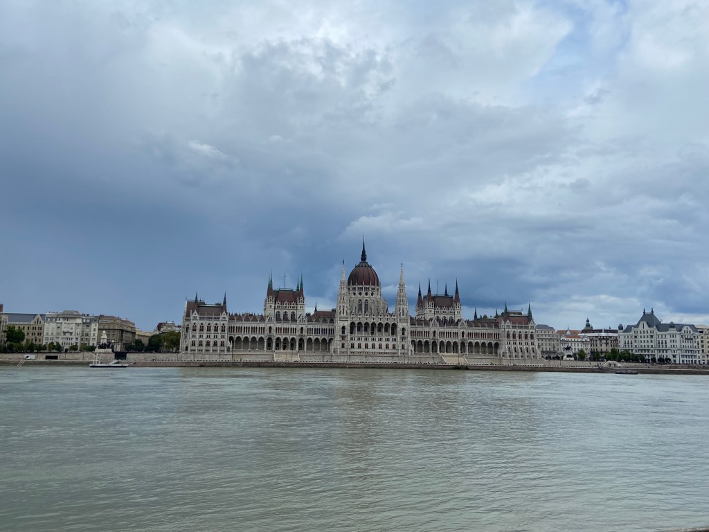 Grand parliamentary building built in a gothic revival style. It has many large archways and windows. The building is right on the banks of the Danube, a wide blue, fast flowing river. 
