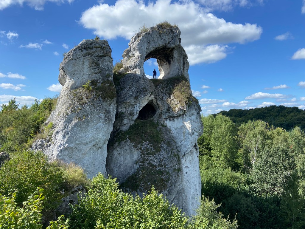 Large white limestone pinnacle set within the forest. On the top of one of the pinnacles there is a large window like opening like the eye of a needle with a person stood in the middle of it.