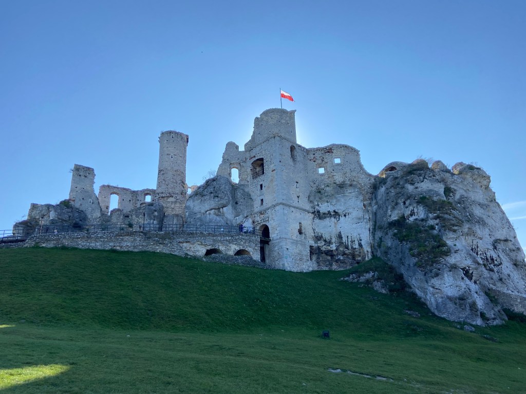 Semi ruined castle fortress built into the rocky sector with a long stone bridge across the grassy hilly. There is a polish crag flying above one of the turrets.