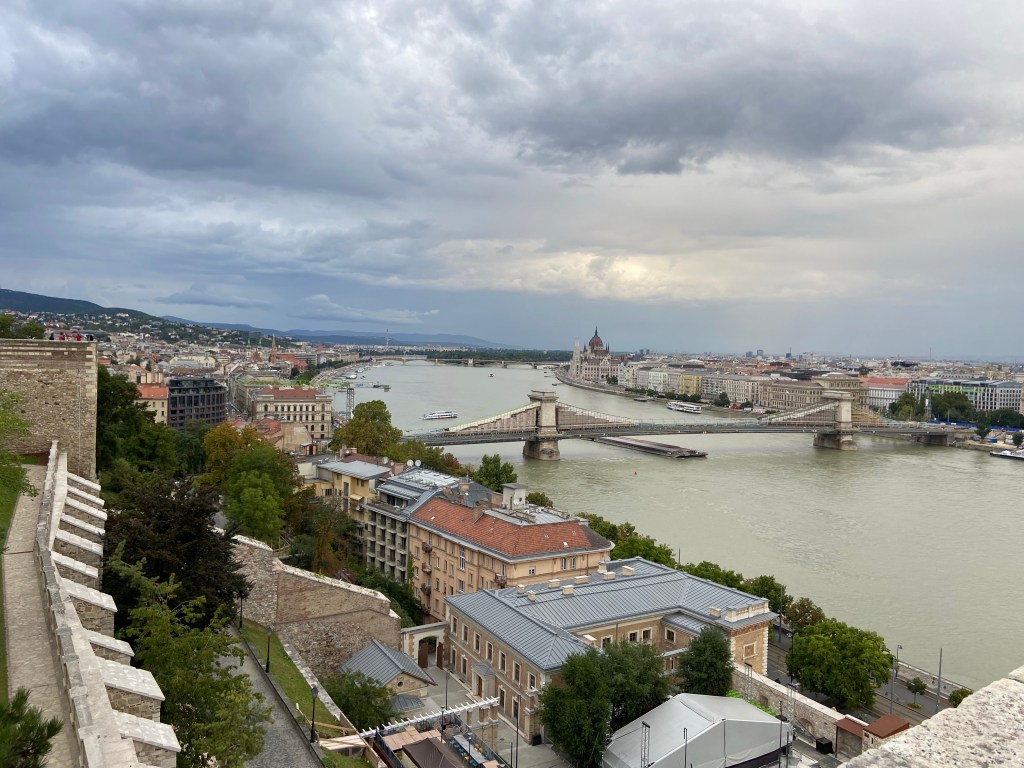 Roof top view looking from the fortress wall over the grand buildings in the city along the famous wide and fast flowing Danube river. There is a flat barge like boat passing under the bridge which connects the two halves of the city. 