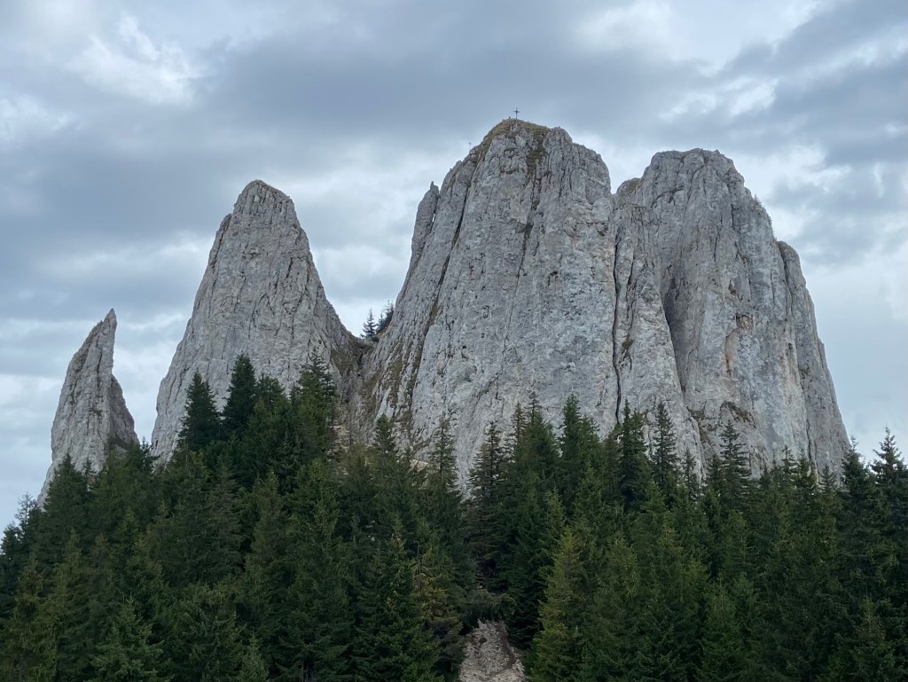 Massif of alpine limestone pinnacles erupting out of the lush green pine forest. At the top of the highest pinnacle of rock is a cross.