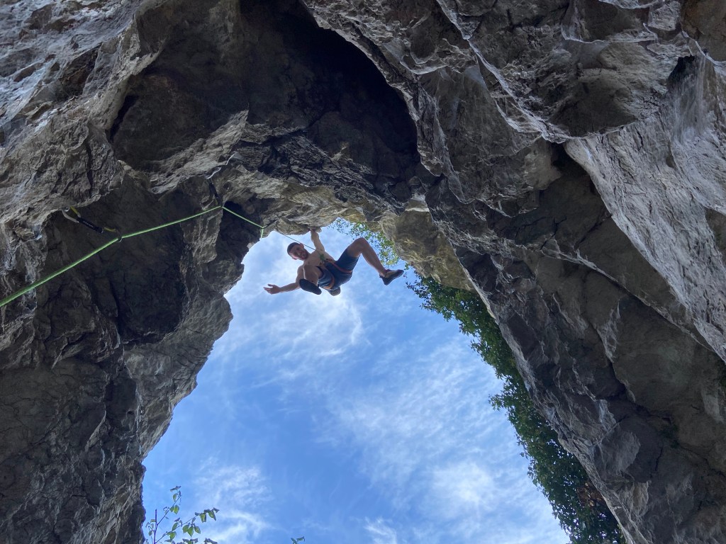 Climber free hanging in space on one arm, on a short but steep climb, looking out towards the sky.