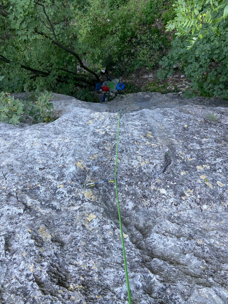 View down towards the belayer stood at the base of the cliff in the forest. The rock is limestone with patches of yellow lichen and a few features. 