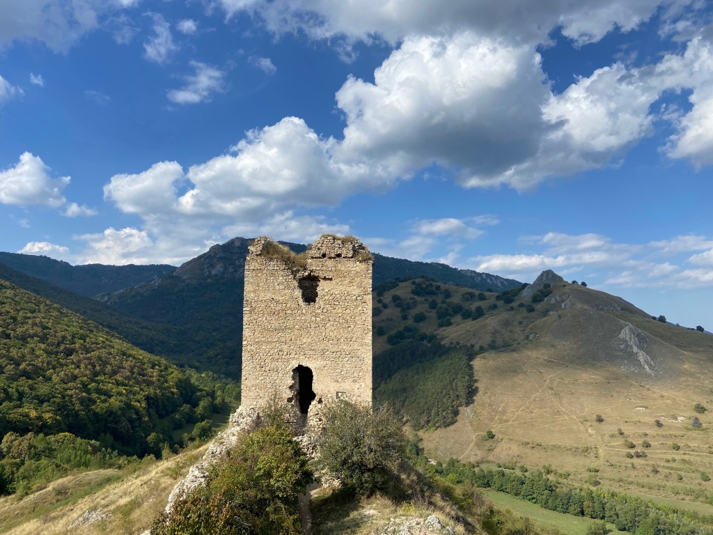 Solitary stone turret from the 13th century, abandoned  and crumbling, set on a hill top overlooking other hills in the distance.  