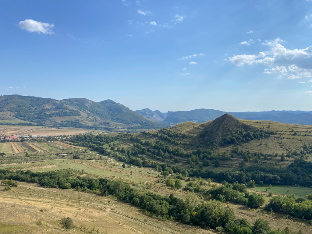 Shallow green valley with mountains and pastured fields. In the foreground is an unusually pointed pyramid like hill. 