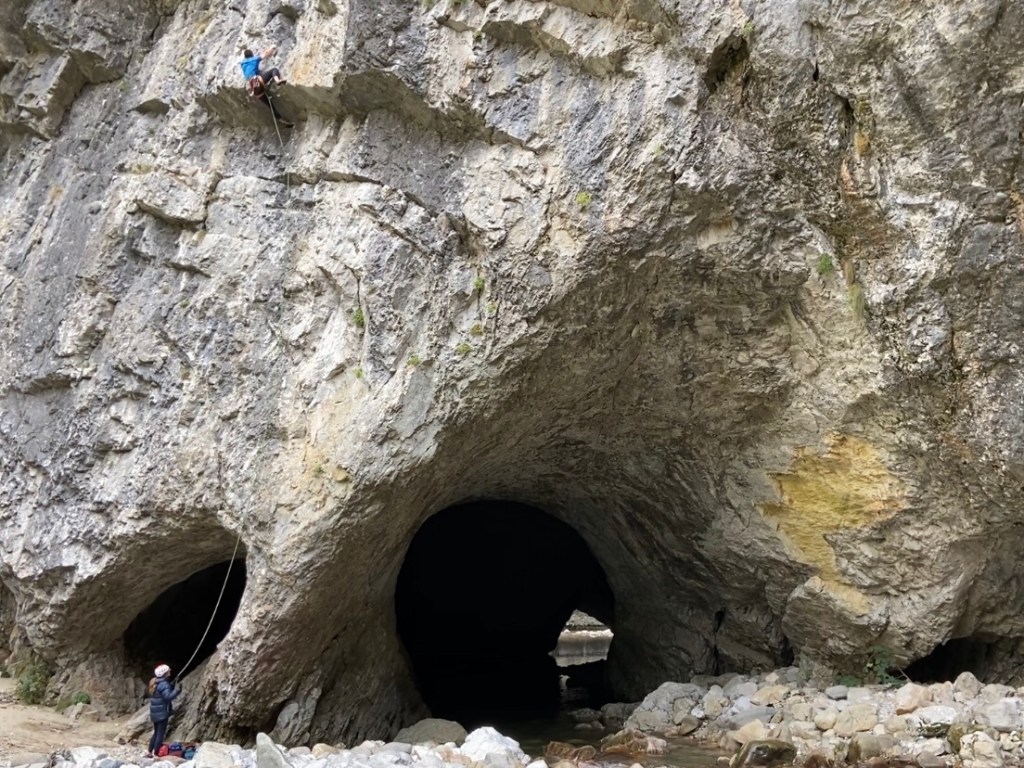 Climber using a high right foot to gain the lip of the roof. Below are two holes, like nostrils, which run the length of the cliff and out to the other side where you can see daylight through. There is a river which runs through the right side. 