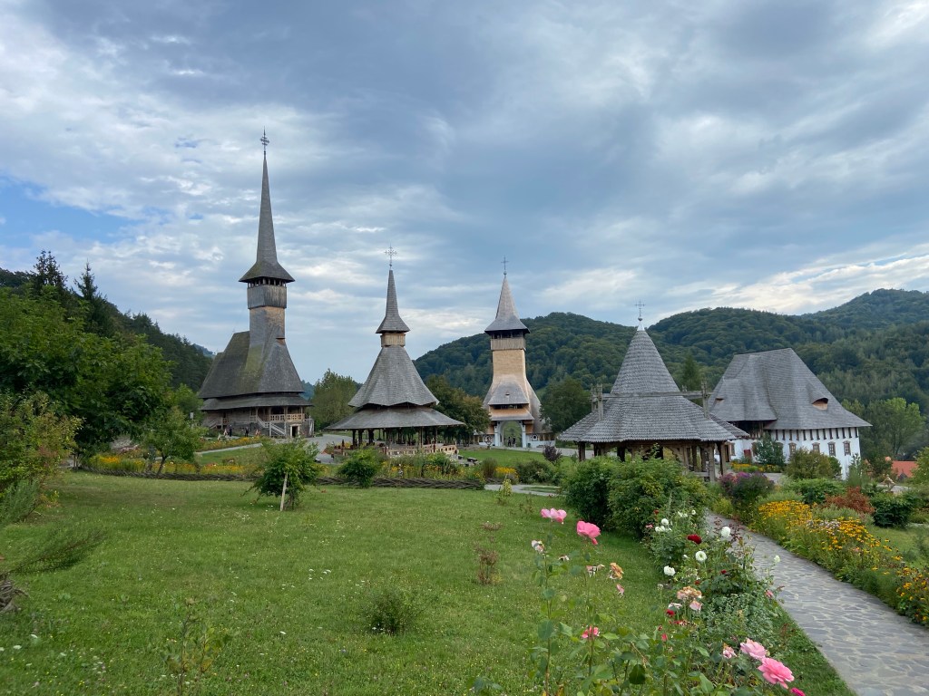 Interesting collection of wooden churches and buildings set within a serene garden filled with flowers. All the buildings have narrow and tall roofs adorned with orthodox crosses on the top  