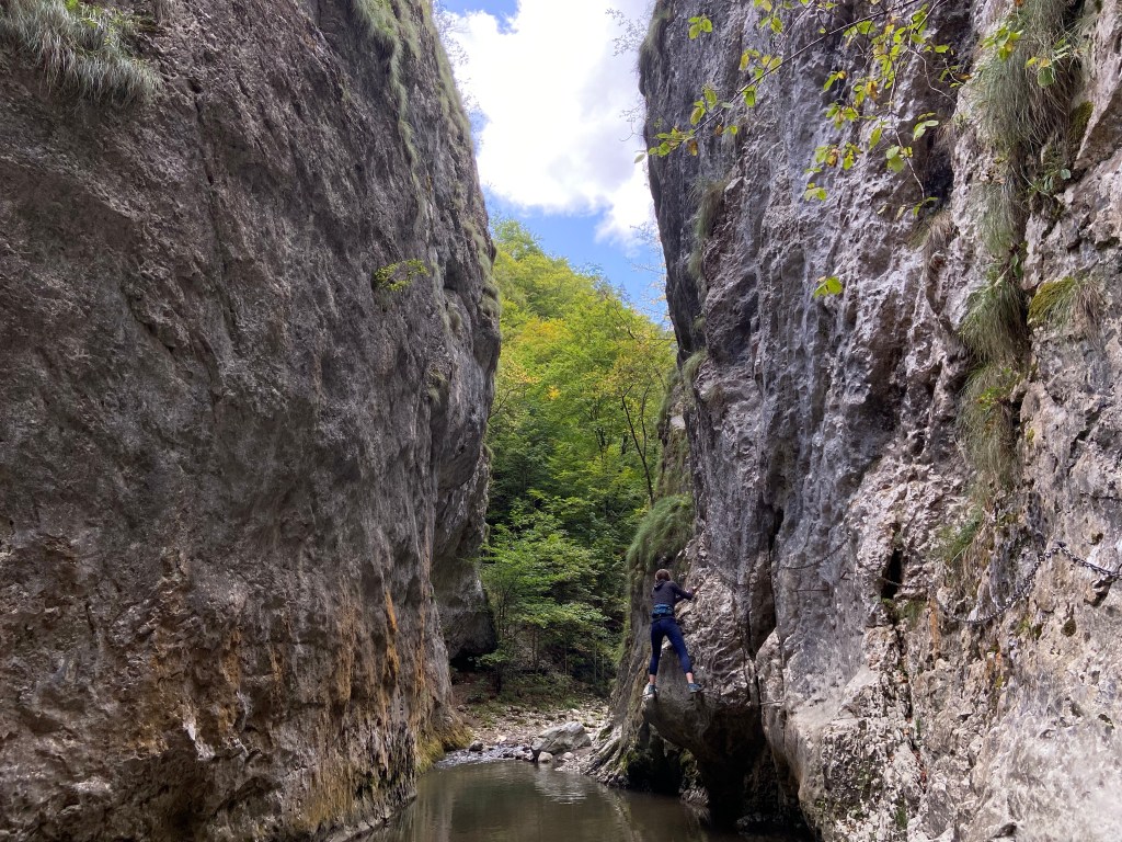 Person using iron cables on the cliff side to navigate a river flowing through a narrow canyon.