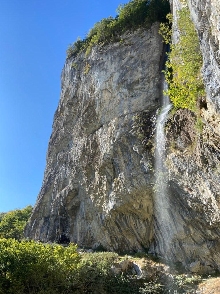 Waterfall falling down a tall limestone cliff which is steep and undercut at the bottom which allows the waterfall to fall in free air. The limestone cliff is white with blue streaks with steep and featured climbing 