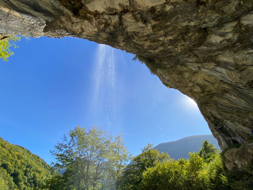 A view from beneath the overhanging cliff with the waterfall flowing in front of the cliff. The view is looking out towards thick forest, more mountains and blue sky. 