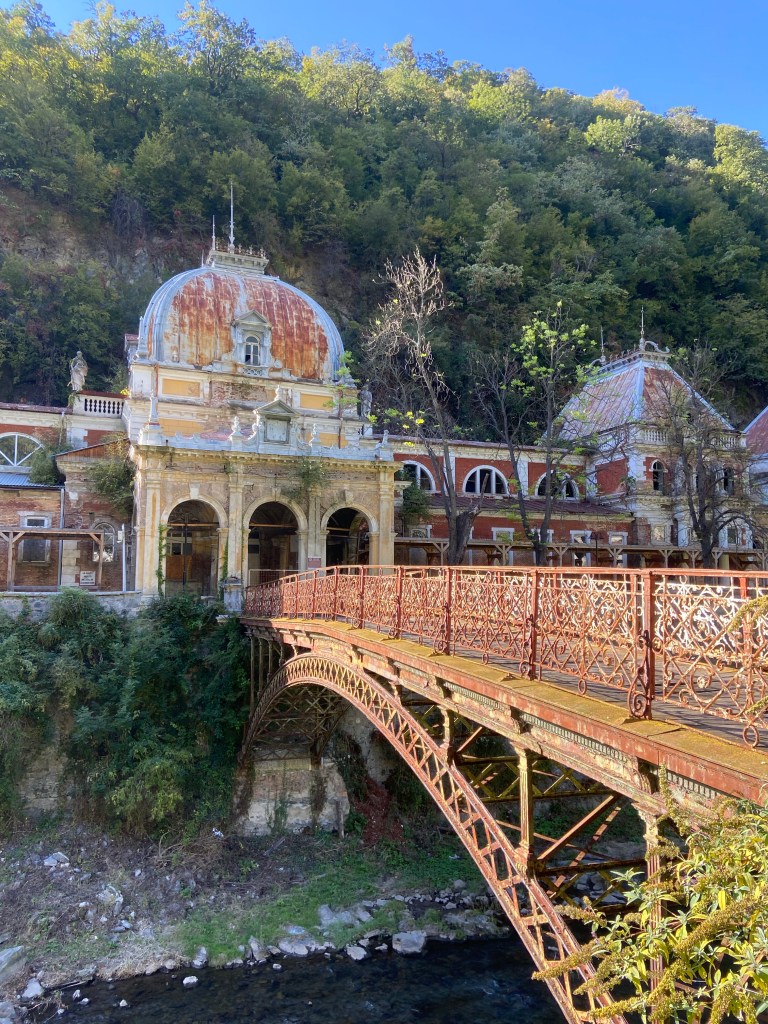 Grand building with domed roof, huge arch ways and multiple wings, abandoned and decaying. The metal work on the roof and on the ornate bridge to cross the river to the building have tuned a bright orange rust colour. 