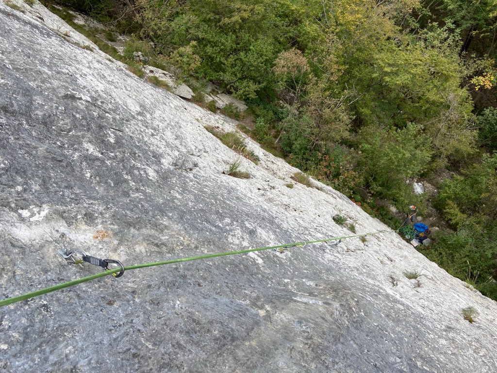 Looking down on a thin and featureless slab towards the belayer in the forest. 