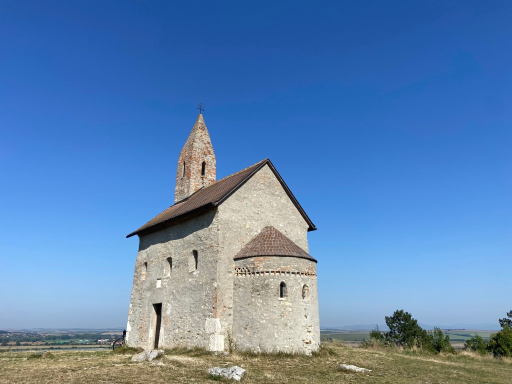 Old stone church set on top of a hill overlooking gently rolling grassy fields into the distance. The church has a spire with a double-barred metal cross on top. 
