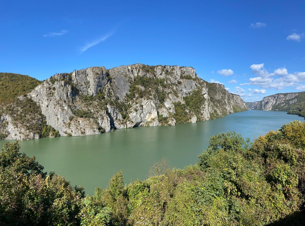 Craggy hillside on the other side of the wide Danube river. The banks of the river forested sides on either side. The river cuts through the valley which forms the border between Romania and Serbia. 