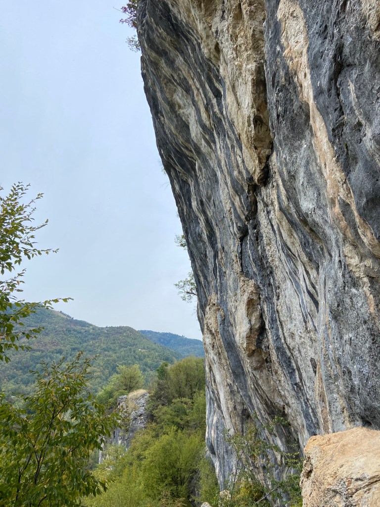 Tall and slightly overhanging limestone wall which is covered in flake features and tufas which have white and blue grey strikes running down the wall. The wall is set with a lush green deciduous forest on rolling hills. 