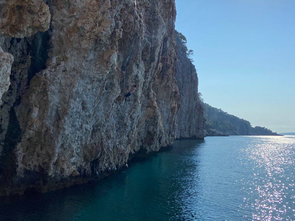 Climber tackling a steep limestone wall high above the water. The limestone cliff extends across the shore line for several hundred meters