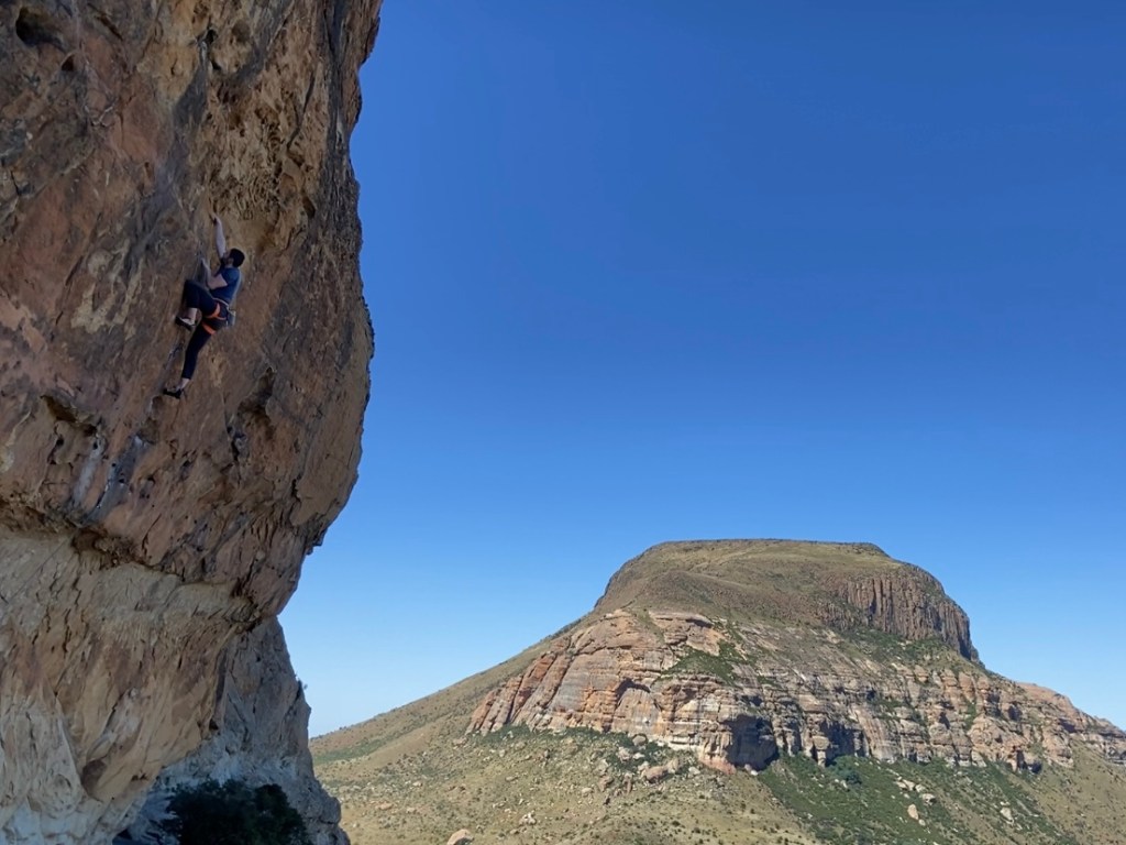 Climber on an overhanging sandstone wall with another sandstone cliff in the background. 