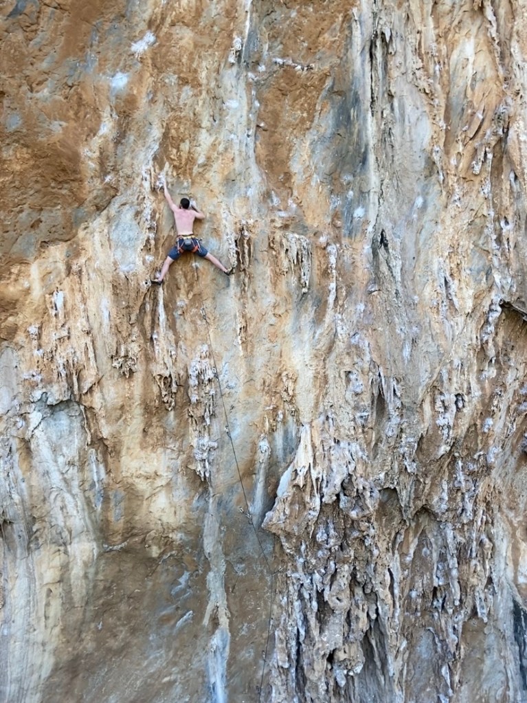 Climber stemming wide across two sets of tufas reaching up high for the next handhold. 