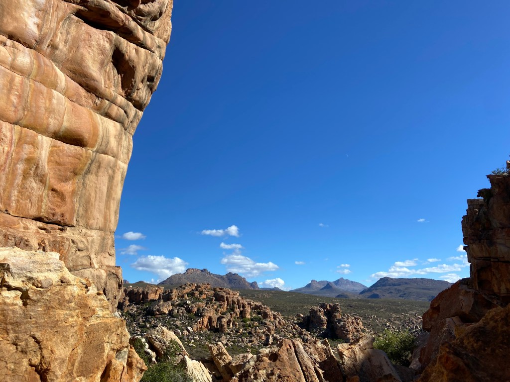 Smooth sandstone wall baked in the heat of the sun, with mountains and boulder fields in the distance. 