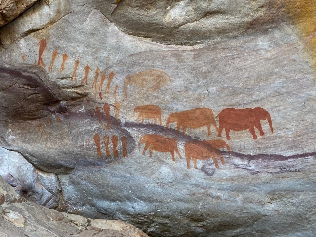 Vibrant orange ancient art work of the San people with and without cloaks and of large elephants.