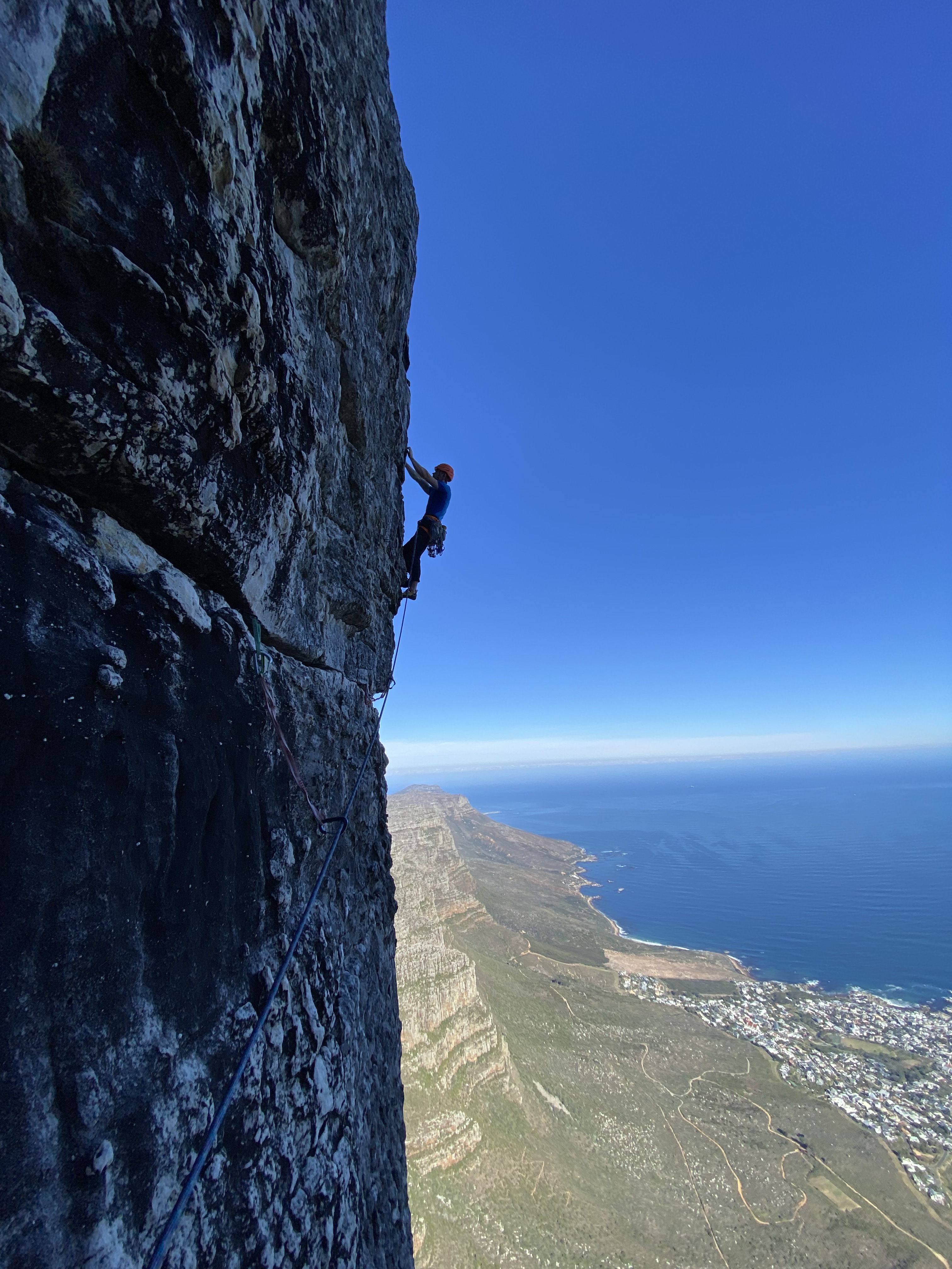 Climber silhouetted against the blue sky and a towering sandstone cliff, with the green mountainside, blue sea and city below