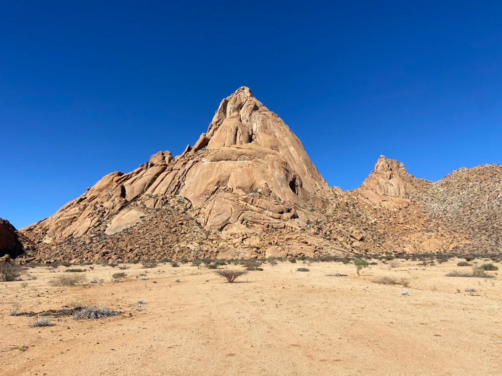 Tall but broken north face of the granite peak of Spitzkoppe. The base of the wall is a large boulder field levelling off to the flat, desert plains.