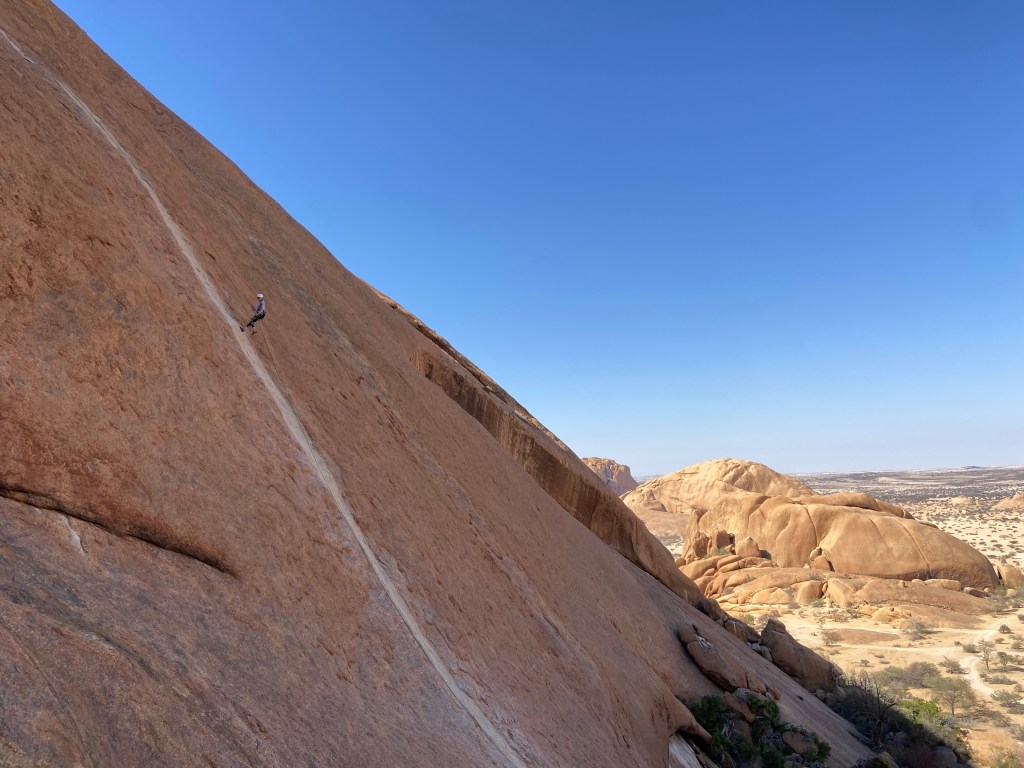 Climber abseiling down a white water worn streak on a large orange granite wall with the desert in the background.