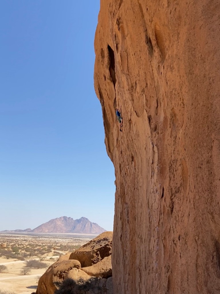 Climber on vertical orange red granite, behind is a desert environment with a huge rocky mountain in the distance.