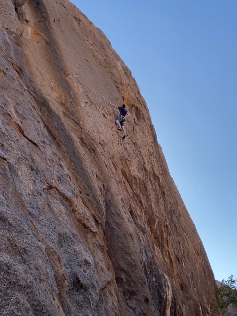 Climber chalking up on a vertical wall climb. The rock is a dark orange with black streaks running down.